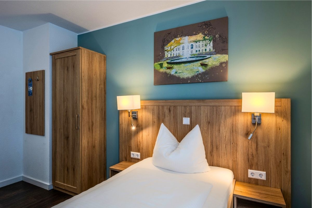 Einzelzimmer im Businesshotel Berlin mit Kleiderschrank, Leseleuchten am Holzkopfteil und Wandbild eines Berliner Schlosses